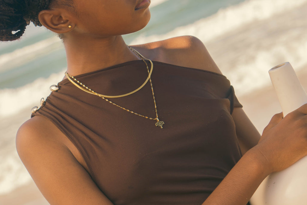 Woman holding a white vase on a beach wearing gold Herringbone and Myrtle Tree Jewellery