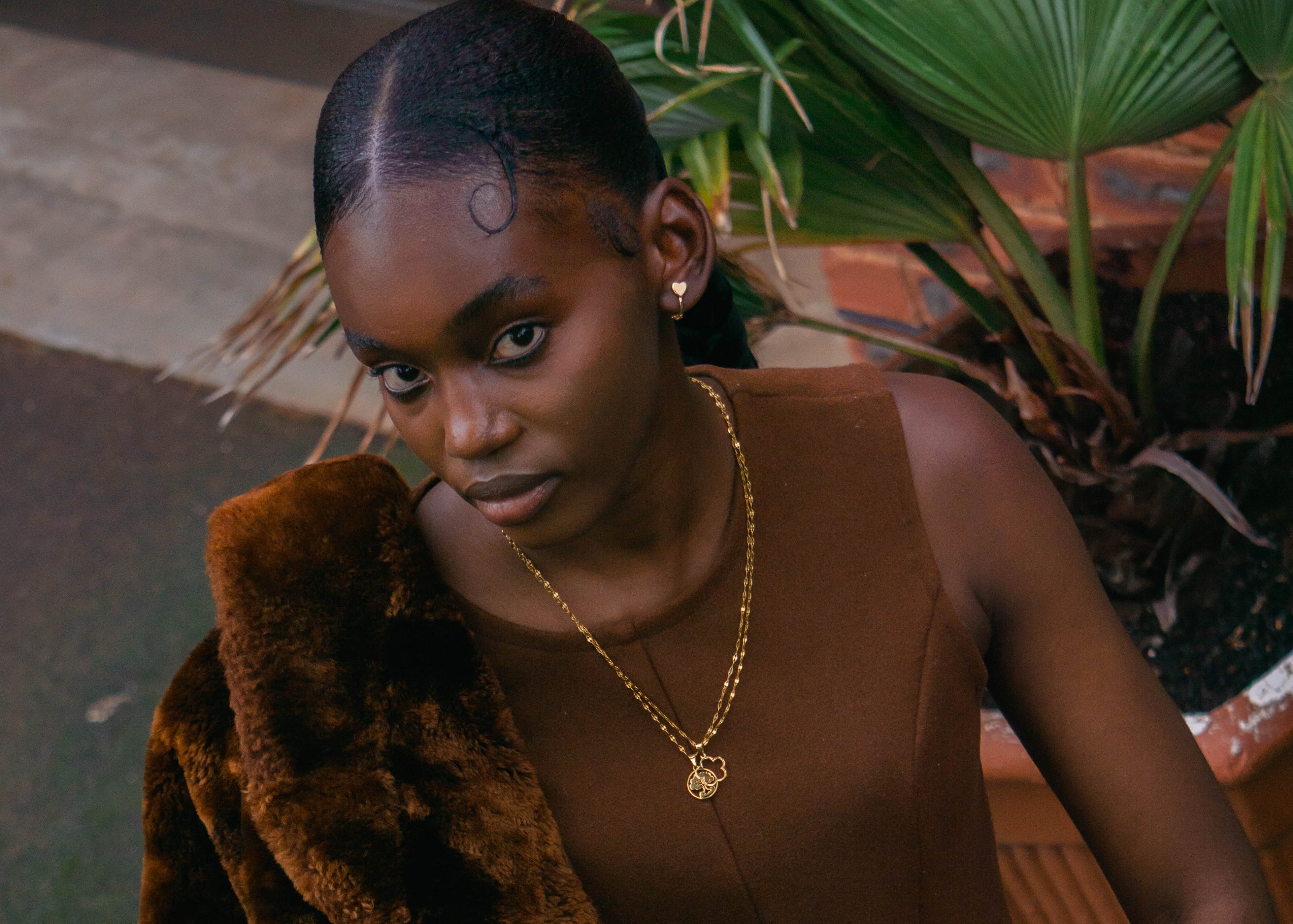 Woman in brown fur coat and dress holding a clutch, standing outdoors with plants in the background